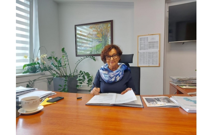 Janina Żagan, Starosta Cieszyński, siedzi przy biurku w gabinecie, wśród dokumentów i kwiatów, z filiżanką herbaty, uśmiechnięta, w okularach i z elegancką apaszką, podpisuje przygotowane pisma.