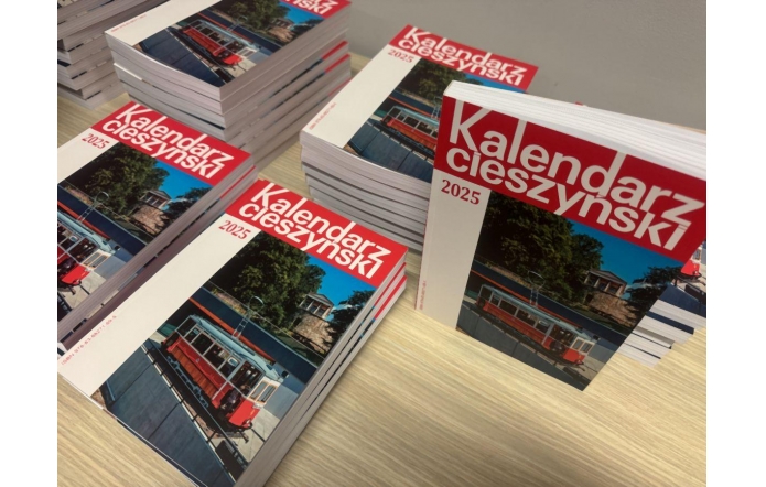 Stosy książek zatytułowanych „Kalendarz Cieszyński 2025” ułożone na jasnym drewnianym blacie. Okładki w odcieniach czerwieni, bieli i błękitu prezentują zdjęcie czerwonego tramwaju na tle budynku otoczonego zielenią. Publikacje są świeżo wydrukowane, równomiernie ułożone w stosy, gotowe do dystrybucji i promocji.