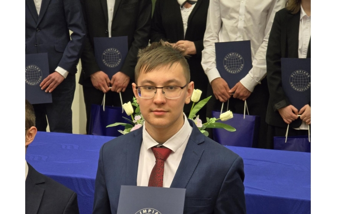 Paweł Dziwoki - uczestnik Olimpiady Geograficznej w garniturze trzyma dyplom na tle grupy innych finalistów. Wszyscy ubrani formalnie, w tle stół z niebieskim obrusem i kwiatami. Widoczne logo „Olimpiada Geograficzna”.