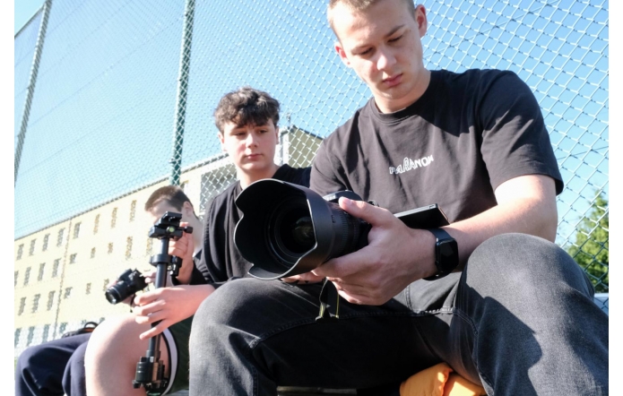 Trzech chłopaków siedzi przy ogrodzeniu boiska, trzymając sprzęt fotograficzny. Jeden z nich skupia się na aparacie z dużym obiektywem, drugi trzyma stabilizator z kamerą, trzeci patrzy z zainteresowaniem.