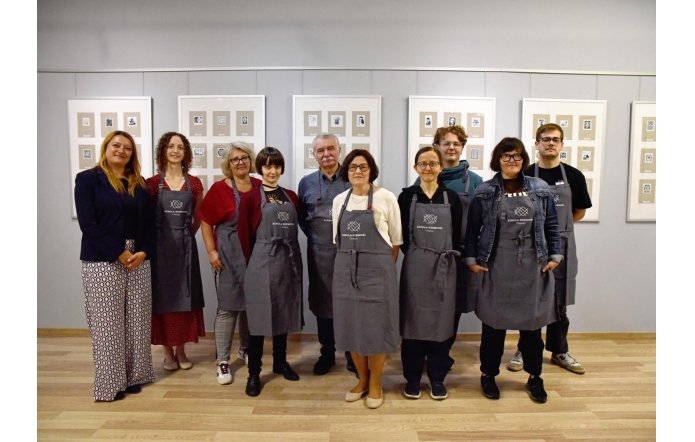 Grupa dziesięciu dorosłych stoi ramię w ramię w jasnej sali wystawowej. Większość ma szare fartuchy z logo „Szkoła Rzemiosł”, po lewej kobieta w granatowej marynarce bez fartucha. W tle równo zawieszone ramy z małymi grafikami, na podłodze jasne deski.