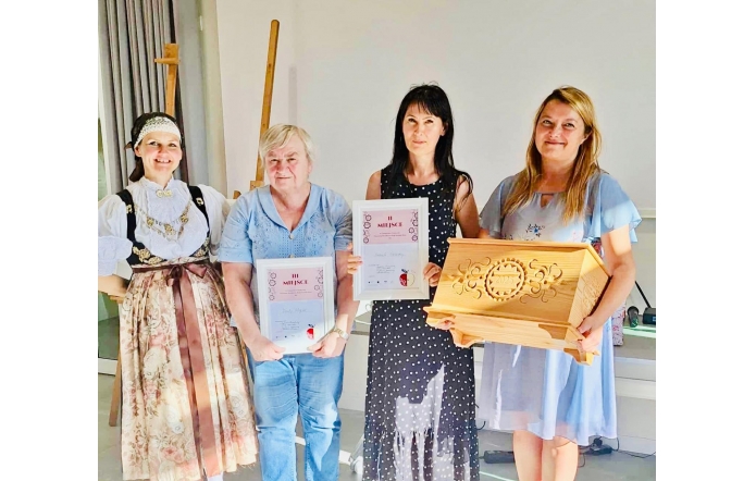 Cztery kobiety pozują w jasnym wnętrzu. Po lewej osoba w bogatym stroju ludowym, obok dwie laureatki trzymają dyplomy z napisami „III MIEJSCE” i „II MIEJSCE”. Po prawej kobieta w błękitnej sukience prezentuje duży, rzeźbiony drewniany kufer z datą 2025. Atmosfera uroczysta i radosna.