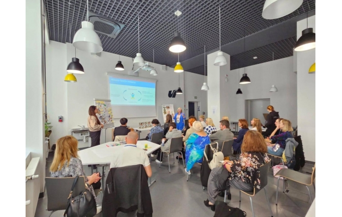 W nowoczesnej sali warsztatowej prowadząca prezentuje slajd przy ekranie.Uczestnicy siedzą przy stołach i na krzesłach, słuchają i notują; na stołach widać markery i materiały. W tle druga prowadząca w niebieskim żakiecie.