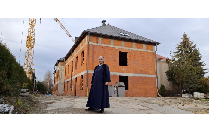 Na tle budowy nowego budynku z czerwonej cegły stoi uśmiechnięta siostra Honorata Klonowska w granatowym habicie z krzyżem na piersi. Po lewej stronie widać żółty dźwig, a wokół placu budowy rozłożone są materiały budowlane. Za budynkiem rosną drzewa iglaste.
