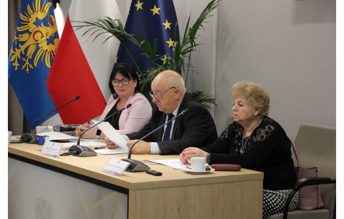 Trzy osoby siedzą przy stole konferencyjnym z mikrofonami; starszy mężczyzna pośrodku czyta dokument, a po jego bokach dwie kobiety uważnie słuchają. W tle znajdują się flagi Polski i Unii Europejskiej oraz roślina.