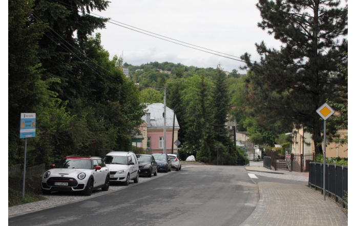 Wąska ulica otoczona zielenią, po lewej szereg zaparkowanych aut, w tym biały Mini z czerwonym dachem. Po prawej ogrodzenia i znak pierwszeństwa, w tle pastelowe domy oraz zalesione wzgórza pod pochmurnym niebem.
