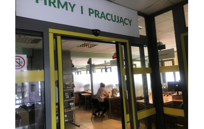 Wejście do biura z napisem „FIRMY I PRACUJĄCY”. Wewnątrz znajdują się stanowiska obsługi, przy jednym z nich siedzi kobieta. Po lewej stronie stoi stojak z ulotkami. Na szklanych drzwiach widoczny znak zakazu używania telefonów.