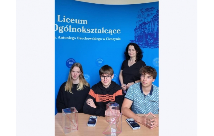 Trzech uczniów i nauczycielka pozują przy stole na tle niebieskiej ścianki z napisem o I LO w Cieszynie. Na stole leżą kalkulatory i trójwymiarowe modele brył geometrycznych z przezroczystego tworzywa.