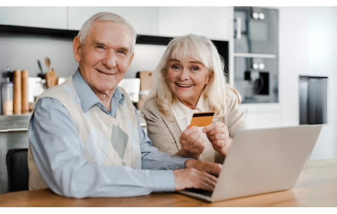 Starsza para siedzi przy stole w nowoczesnej kuchni, uśmiechnięta i zadowolona, korzysta z laptopa. Kobieta trzyma w ręku kartę płatniczą, a mężczyzna kładzie dłoń na klawiaturze. Atmosfera jest serdeczna i pełna spokoju.