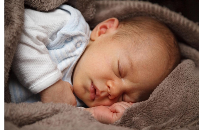 Śpiący niemowlak otulony miękkim, brązowym kocykiem, twarz spokojna, usta lekko rozchylone. Ubrany w jasne body, malutka dłoń zaciśnięta tuż przy policzku.
