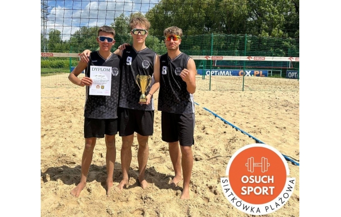 Trzech chłopców w sportowych strojach stoi boso na piaskowym boisku do siatkówki plażowej. Jeden trzyma dyplom, drugi puchar. W tle siatka, banery sponsorów i drzewa. W rogu grafika z napisem „Osuch Sport”.