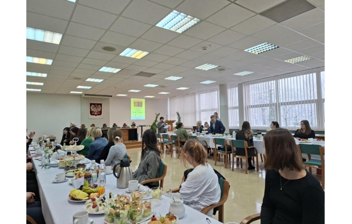 Na zdjęciu widać salę konferencyjną, w której przy długich stołach siedzą uczestnicy spotkania. Na stołach ustawione są przekąski i napoje. Z przodu sali jedna osoba przemawia przy mównicy, a obok niej stoi prowadzący. W tle widoczny jest ekran z prezentacją oraz godło Polski.