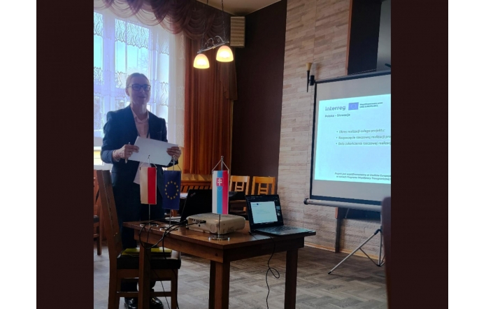 Kobieta stojąca przy stole trzyma dokument. Na stole znajduje się projektor, laptop oraz małe flagi Polski, Unii Europejskiej i Słowacji. Obok ustawiony jest ekran z wyświetloną prezentacją programu Interreg Polska-Słowacja. W tle widoczne są krzesła, okno z zasłonami i oświetlenie sufitowe.