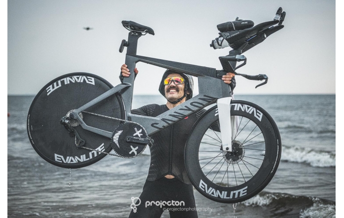 Mężczyzna w stroju sportowym i kasku unosi nad głową rower, stojąc w płytkiej wodzie przy brzegu morza. Ma na sobie sportowe okulary, a na twarzy widoczny jest uśmiech. W tle rozciąga się morze i pochmurne niebo.