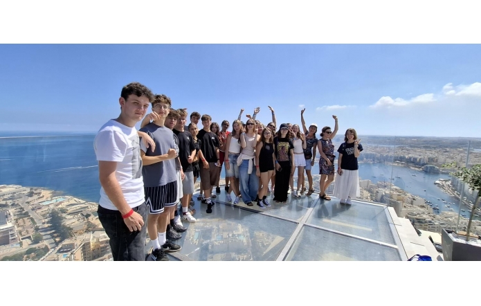 Grupa nastolatków i dorosłych stoi na szklanym tarasie widokowym wysoko nad nadmorskim miastem. Słoneczny dzień, błękitne niebo i rozległe morze z zatoką oraz łódkami w dole. Wszyscy uśmiechnięci, część macha do aparatu, luźne, letnie  stroje.