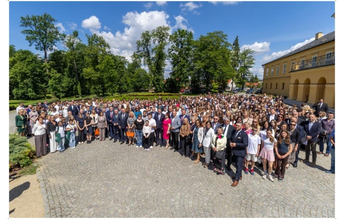 Uczestnicy wydarzenia pozują do wspólnego zdjęcia na dziedzińcu przed budynkiem. Na fotografii widoczna jest liczna grupa osób zgromadzonych na otwartej przestrzeni. W tle znajdują się drzewa, park oraz zabudowania.