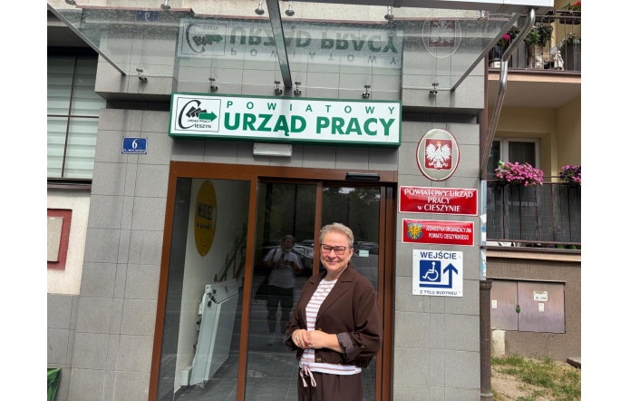 Uśmiechnięta Anna Stefaniak-Bacza Dyrektor Powiatowego Urzędu Pracy w Cieszynie stoi przed wejściem do szarego budynku. Nad drzwiami widoczny jest duży zielony napis „Powiatowy Urząd Pracy”, a obok tablice z godłem Polski, herbem Powiatu Cieszyńskiego oraz informacją o wejściu dla osób z niepełnosprawnościami.