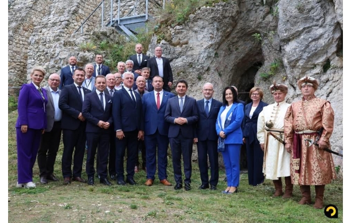 Grupa kobiet i mężczyzn stoi przed skalnymi ruinami. Większość osób ma na sobie garnitury, a po prawej stronie widoczni są dwaj mężczyźni w historycznych strojach z nakryciami głowy. W tle znajdują się kamienne ściany, skały oraz metalowe schody.