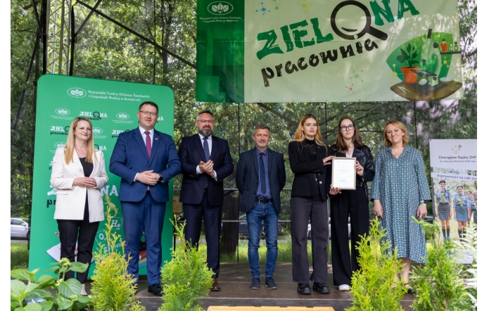 Grupa uśmiechniętych osób stoi na scenie pod banerem „Zielona Pracownia”. Wśród nich są przedstawiciele instytucji i szkół, jedna z kobiet trzyma dyplom. W tle zieleń drzew i dekoracje z roślinami na pierwszym planie.