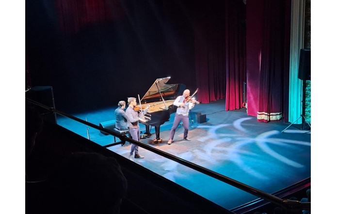 Na scenie występuje trzech muzyków: dwóch skrzypków stoi na przodzie i gra z energią, a za nimi pianista siedzi przy dużym, otwartym fortepianie. Artyści są oświetleni delikatnymi reflektorami, które tworzą jasne wzory na podłodze.