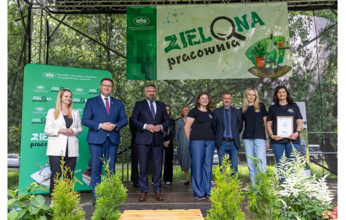 Grupa uśmiechniętych osób stoi na scenie pod banerem „Zielona Pracownia”. Wśród nich są przedstawiciele instytucji i szkół, jedna z kobiet trzyma dyplom. W tle zieleń drzew i dekoracje z roślinami na pierwszym planie.