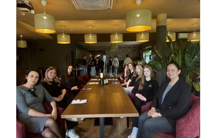 Grupa uczniów w eleganckich strojach hotelarskich ćwiczy przy recepcji. W tle widoczna jest tablica z nazwą obiektu, a atmosfera spotkania jest pełna zaangażowania i uśmiechów.