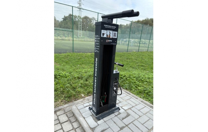 Czarna samoobsługowa stacja naprawy rowerów z wiszącymi narzędziami i pompką, ustawiona na kostce brukowej przy ogrodzonym boisku, wokół trawa.