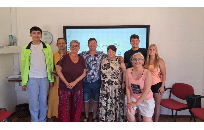 Grupowe zdjęcie dziewięciu osób stojących  w jasnym pomieszczeniu szkoleniowym. W tle widać duży ekran, a obok ścienny zegar. Osoby stoją blisko siebie, uśmiechają się do aparatu, część ma na sobie letnie, kolorowe ubrania. Wokół znajdują się czerwone krzesła i półki z materiałami.