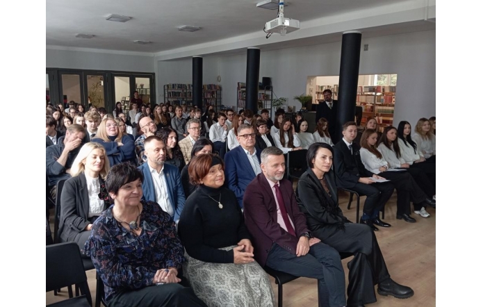 Sala wypełniona siedzącymi ludźmi, głównie dorosłymi i młodzieżową, ustawionymi w rzędach. Wszyscy są elegancko ubrani i skierowani w jedną stronę. W tle znajdują się regały z książkamioraz kolumny.