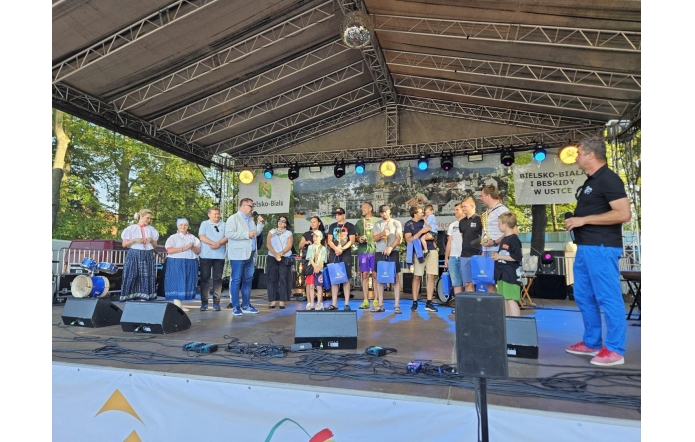 Grupa osób stoi na scenie plenerowej – część w strojach regionalnych, część w sportowych ubraniach lub casualu. Trzymają torby z upominkami. W tle baner z napisem „Bielsko-Biała i Beskidy w Ustce”.