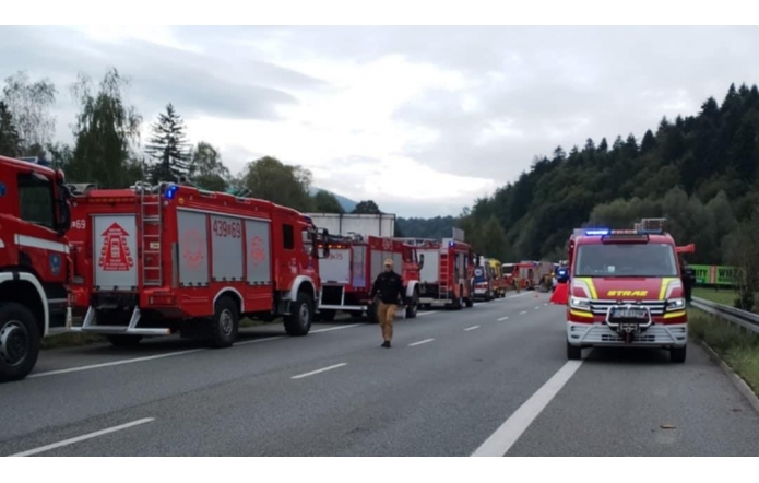 Szereg czerwonych wozów strażackich i karetek stoi na zablokowanej drodze ekspresowej, po pasie awaryjnym stoi bus straży z włączonymi światłami. Między pojazdami idą ratownicy, a w tle widać drzewa i pagórki pod pochmurnym niebem.