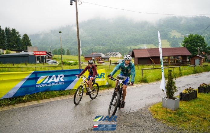 Dwójka rowerzystów na rowerach górskich, jadących po mokrej szosie.W tle zamglony i zalesiony stok. 